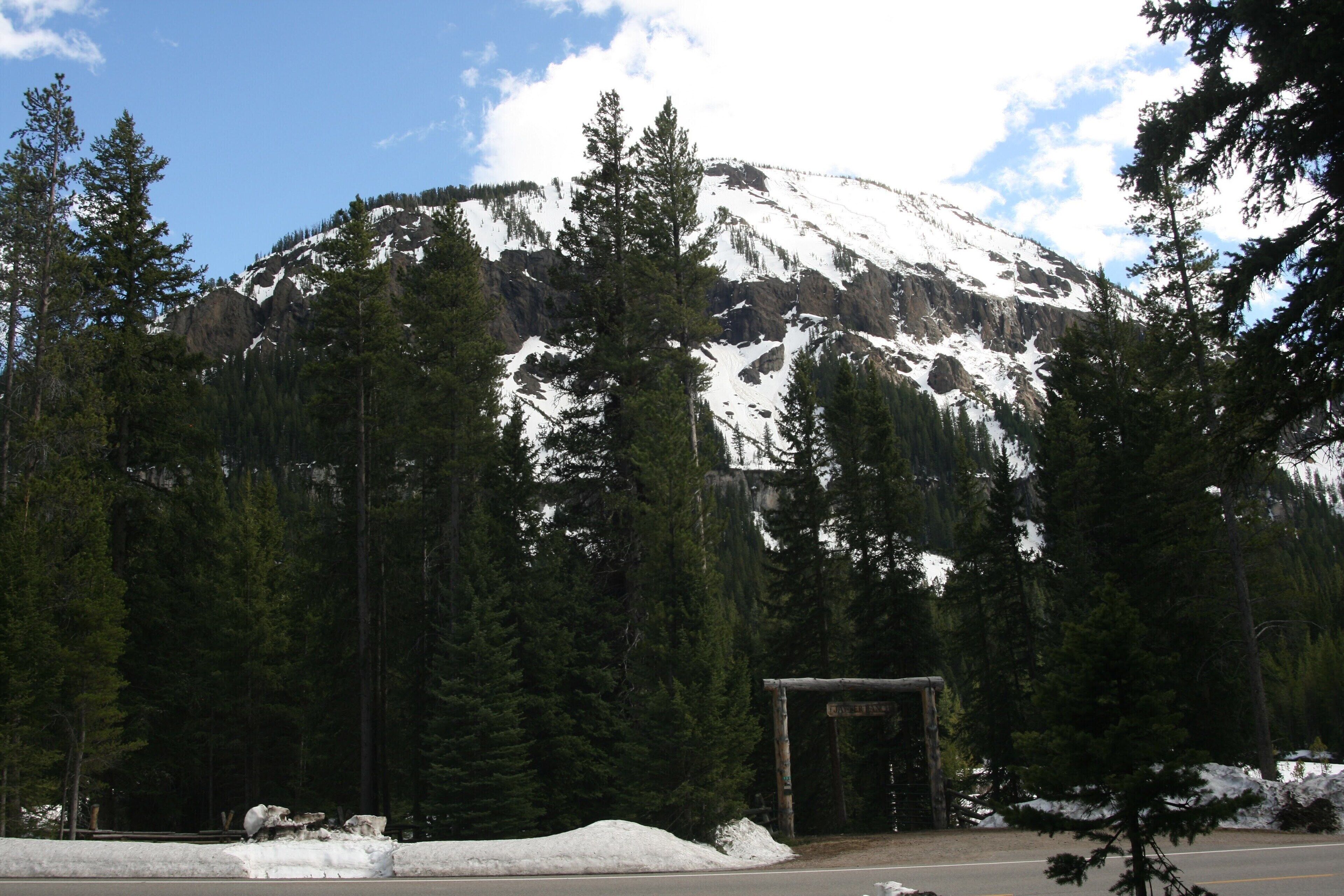 A mountain with snow-covered peaks, dense forest, and a wooden structure in the foreground.