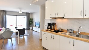Une cuisine moderne avec des meubles blancs, un évier et une machine à café. On y trouve un coin repas avec une table et des chaises, ainsi qu’une grande fenêtre avec des rideaux.