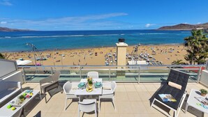 Un balcon avec une table dressée pour deux personnes, donnant sur une plage parsemée de parasols et animée par la présence de nombreux vacanciers.