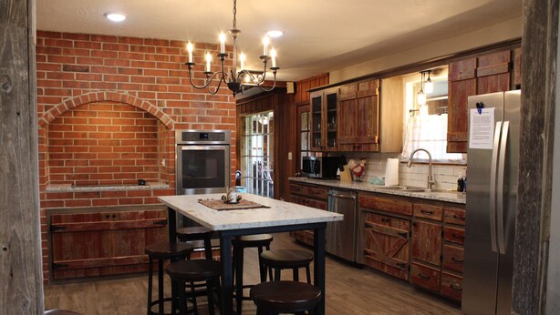 A kitchen with a brick wall, wooden cabinets, a stainless steel refrigerator, and a central island with stools. - HaT Ranch Lodge. Perfect for a large group or multiple families. NO CLEANING FEE (Ponca City)