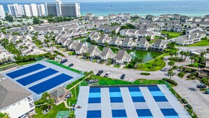 Una zona residencial con varias casas de dos pisos, una gran cancha de tenis azul y un campo deportivo verde más pequeño.