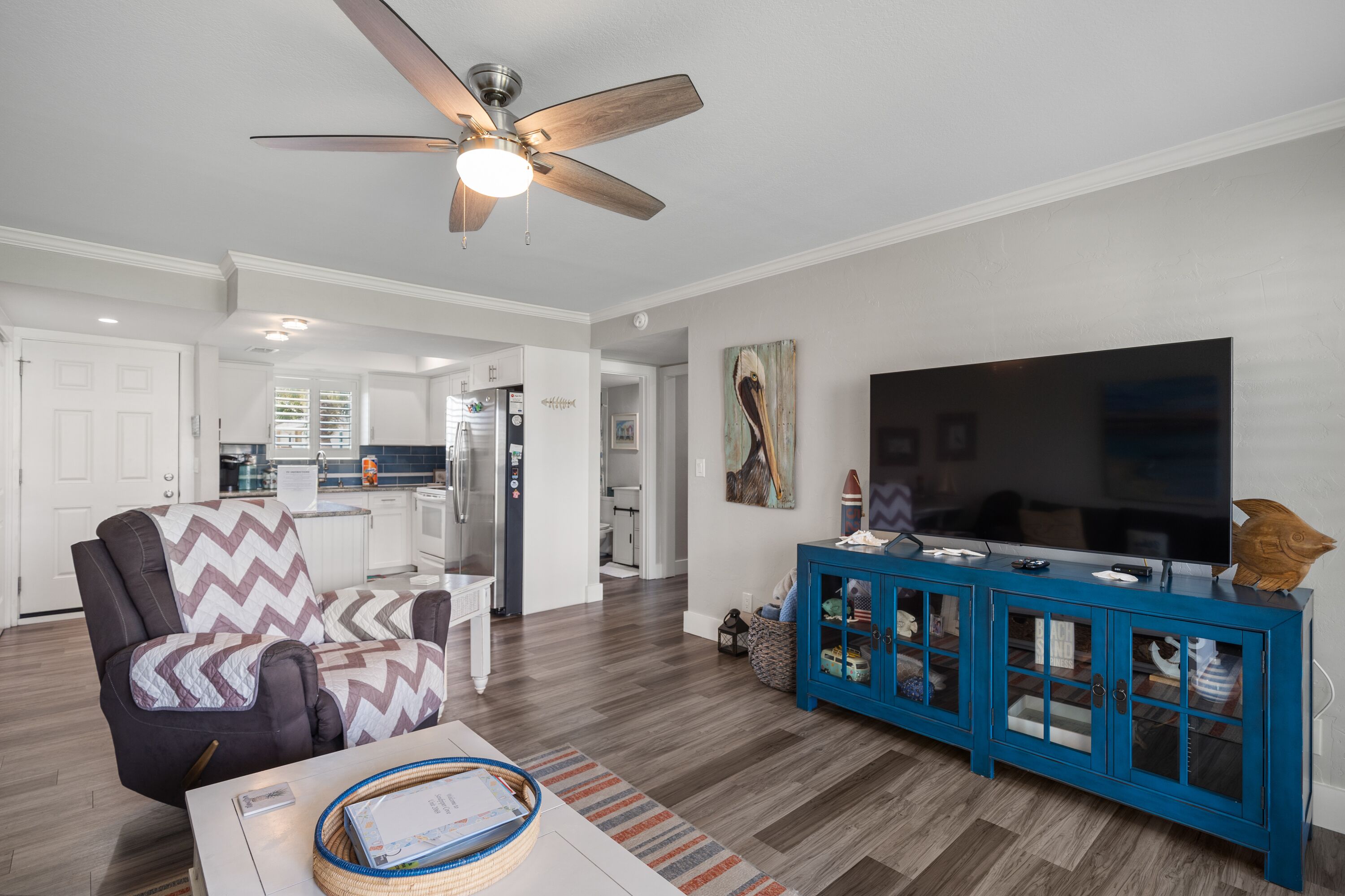 A modern living room with a ceiling fan, a large flat-screen TV, a blue cabinet with glass doors, and a comfortable armchair.