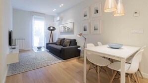A modern living room with a sofa, a dining table with chairs, a TV, and a large window. - Viena | The Rentals Collection (San Sebastián)