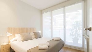 A bedroom with a bed, bedside table, lamp, and a large window with blinds. - Viena | The Rentals Collection (San Sebastián)