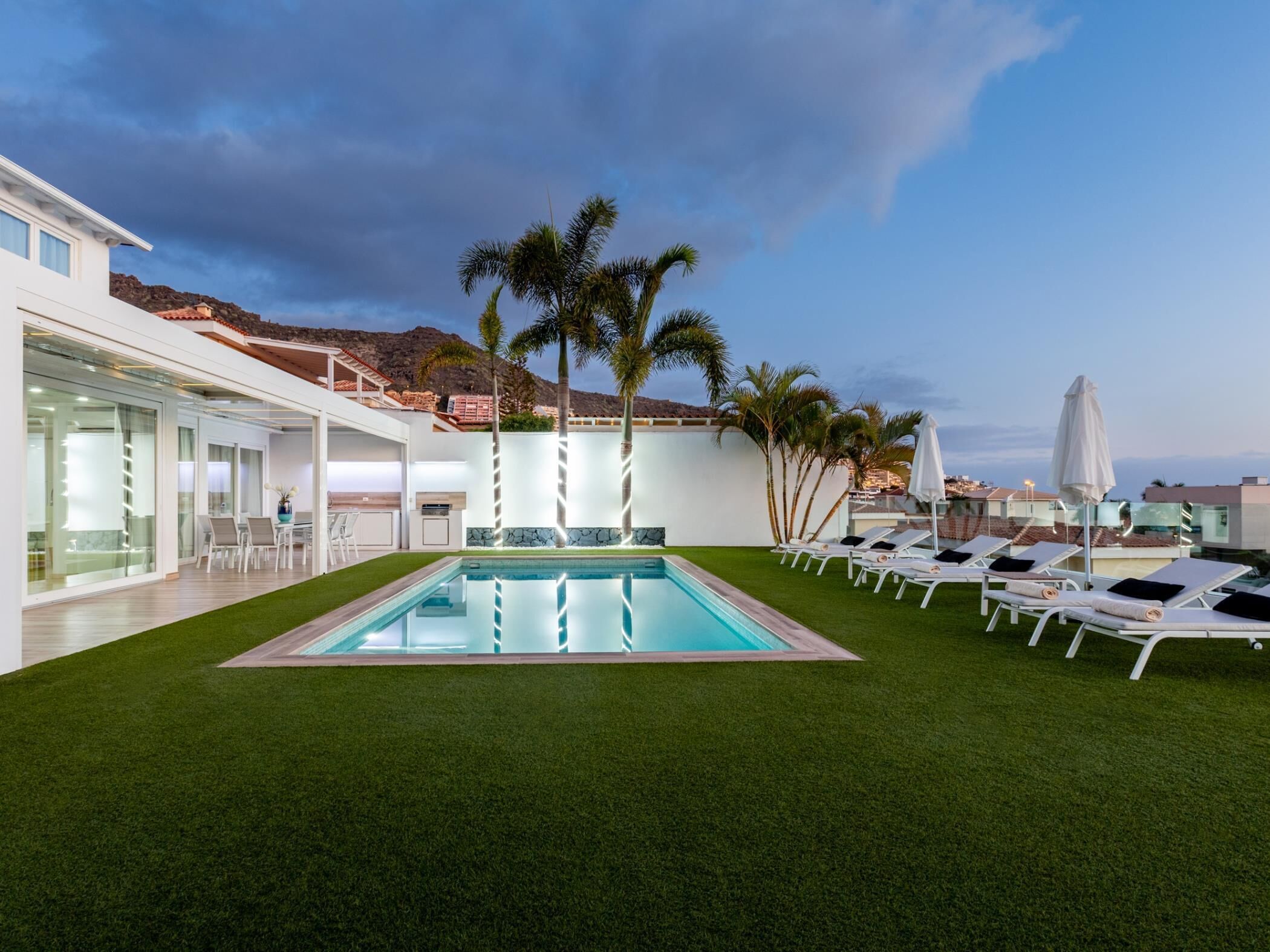 A modern outdoor pool area with a rectangular pool, surrounded by artificial grass, and equipped with sun loungers and umbrellas.