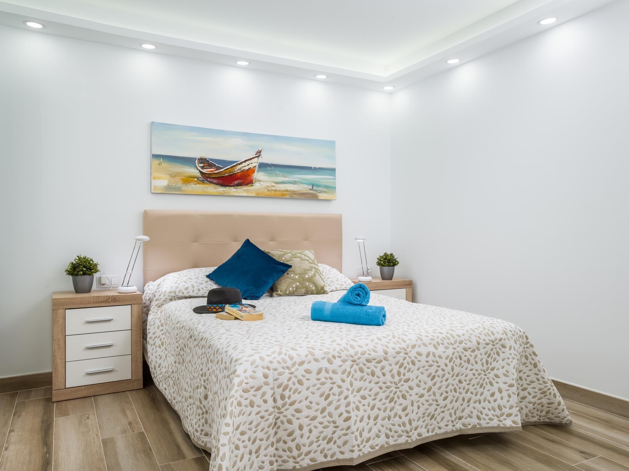 A bedroom with a bed, a nightstand, a lamp, a potted plant, and a painting of a boat on the wall.