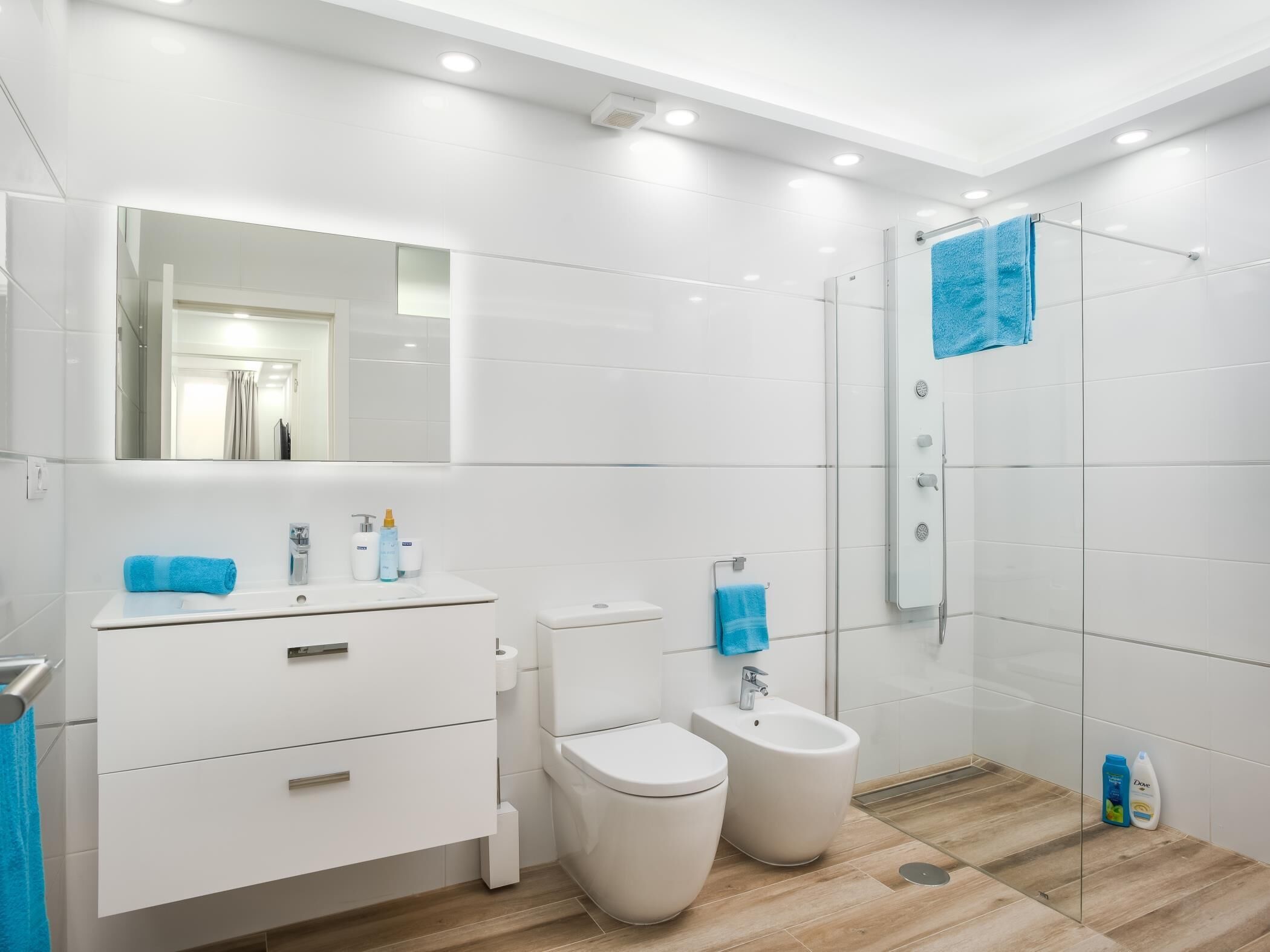 A modern bathroom with white tiles, a large mirror, a sink with toiletries, a toilet, a bidet, and a glass shower enclosure.