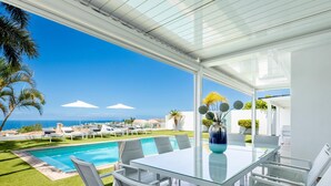 Un área de comedor moderna al aire libre con una mesa y sillas de vidrio, con vistas a una piscina y al océano.