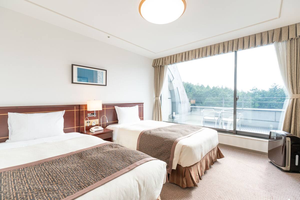 A hotel room with two beds, a TV, a window with a view of trees, and a balcony.