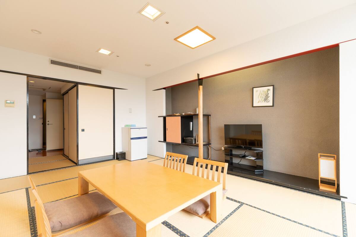 A traditional Japanese room with tatami flooring, a wooden table with cushions, a television, and a sliding door leading to another room.