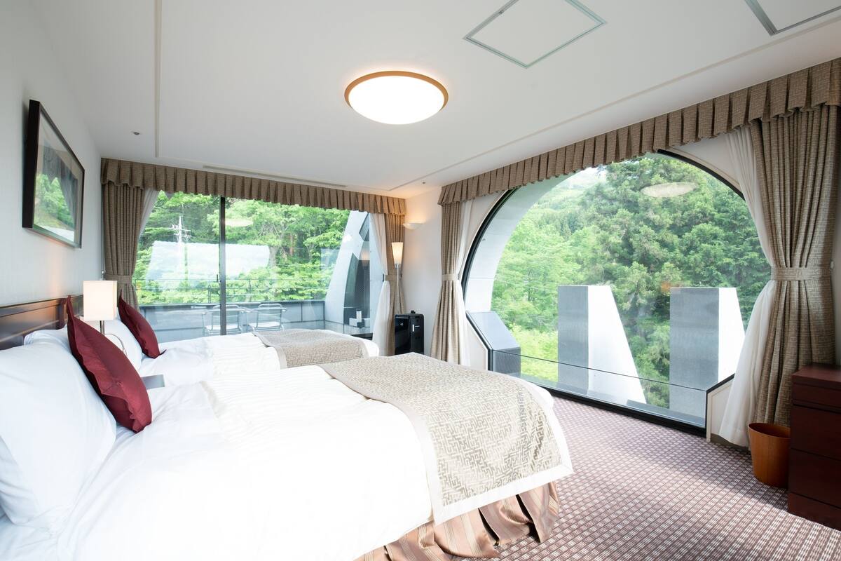 A hotel room with a large bed, two bedside tables, a nightstand, a large window with a view of greenery, and a ceiling light.