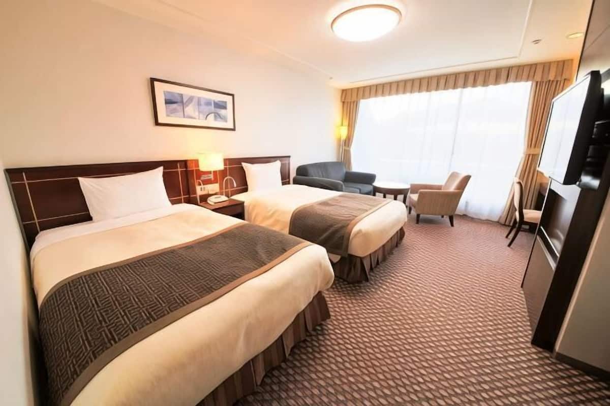 A hotel room with two beds, a desk, a chair, a TV, and a window with curtains.