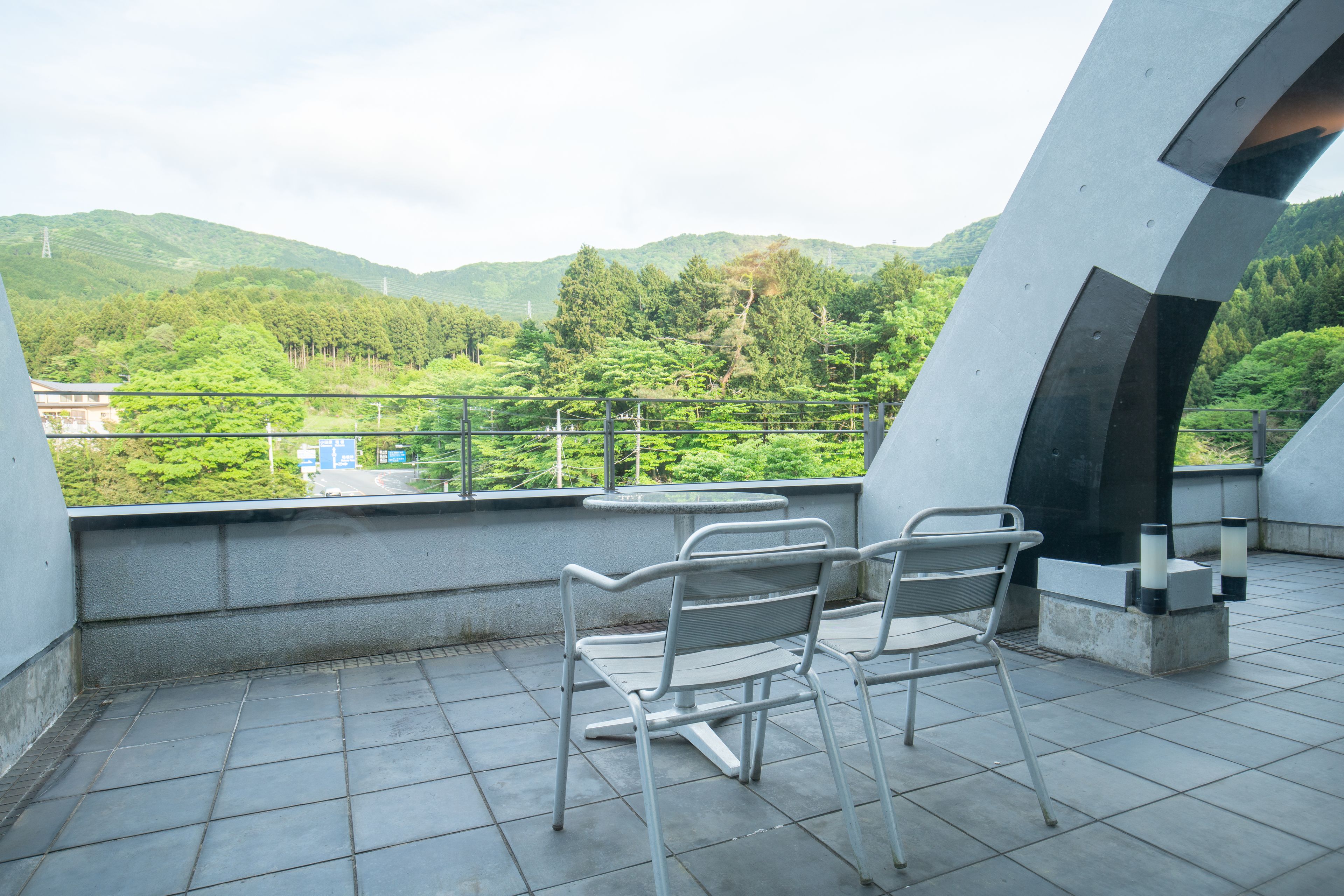 A balcony with two metal chairs, a small table, and a view of green hills and trees.