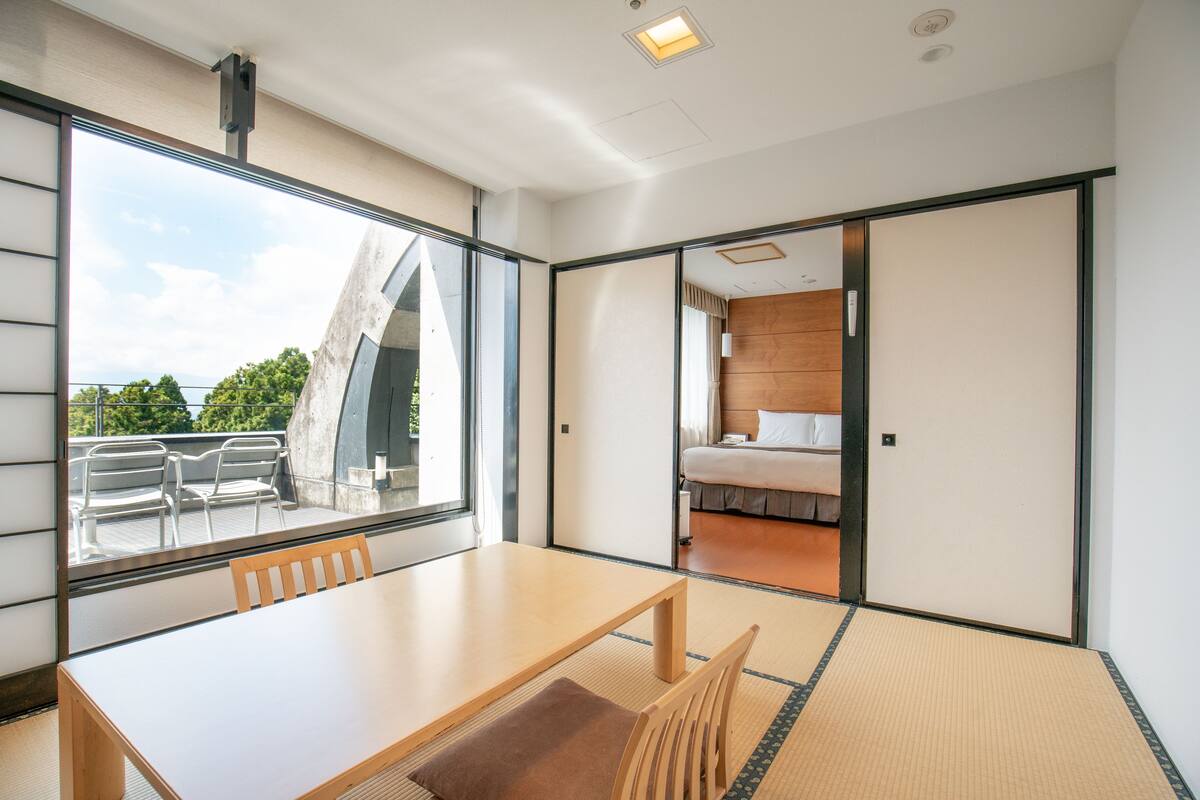A modern hotel room with a large window, a wooden table, and a bed.