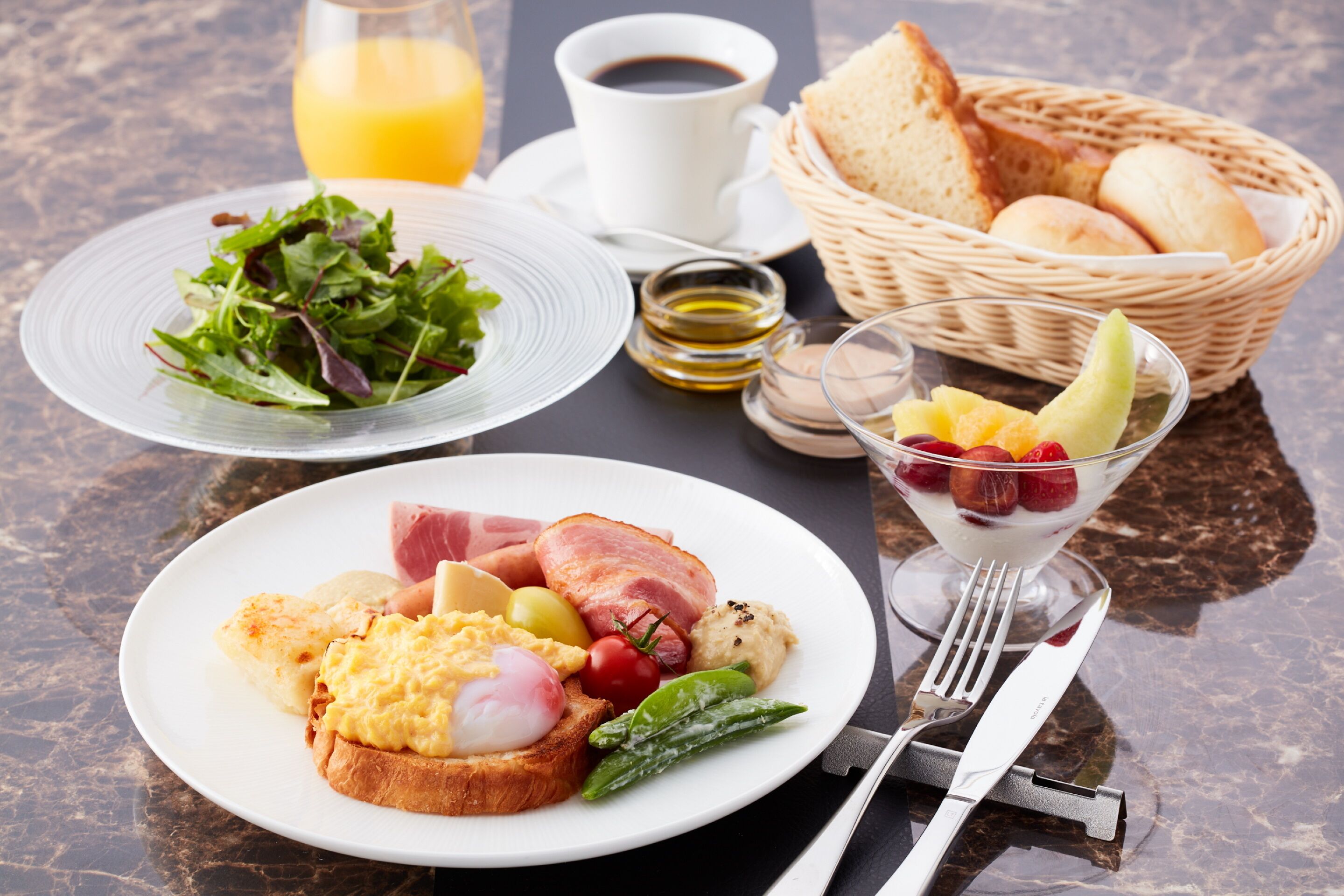 A breakfast setting with toast, eggs, ham, and a salad on a plate, a glass of orange juice, a cup of coffee, and a basket of bread on a marble table.