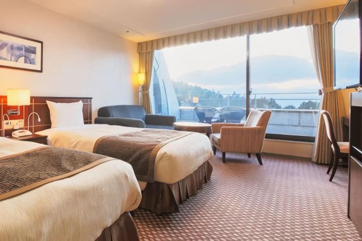 Habitación de hotel con dos camas, un escritorio, una silla, un televisor y un ventanal con vistas.