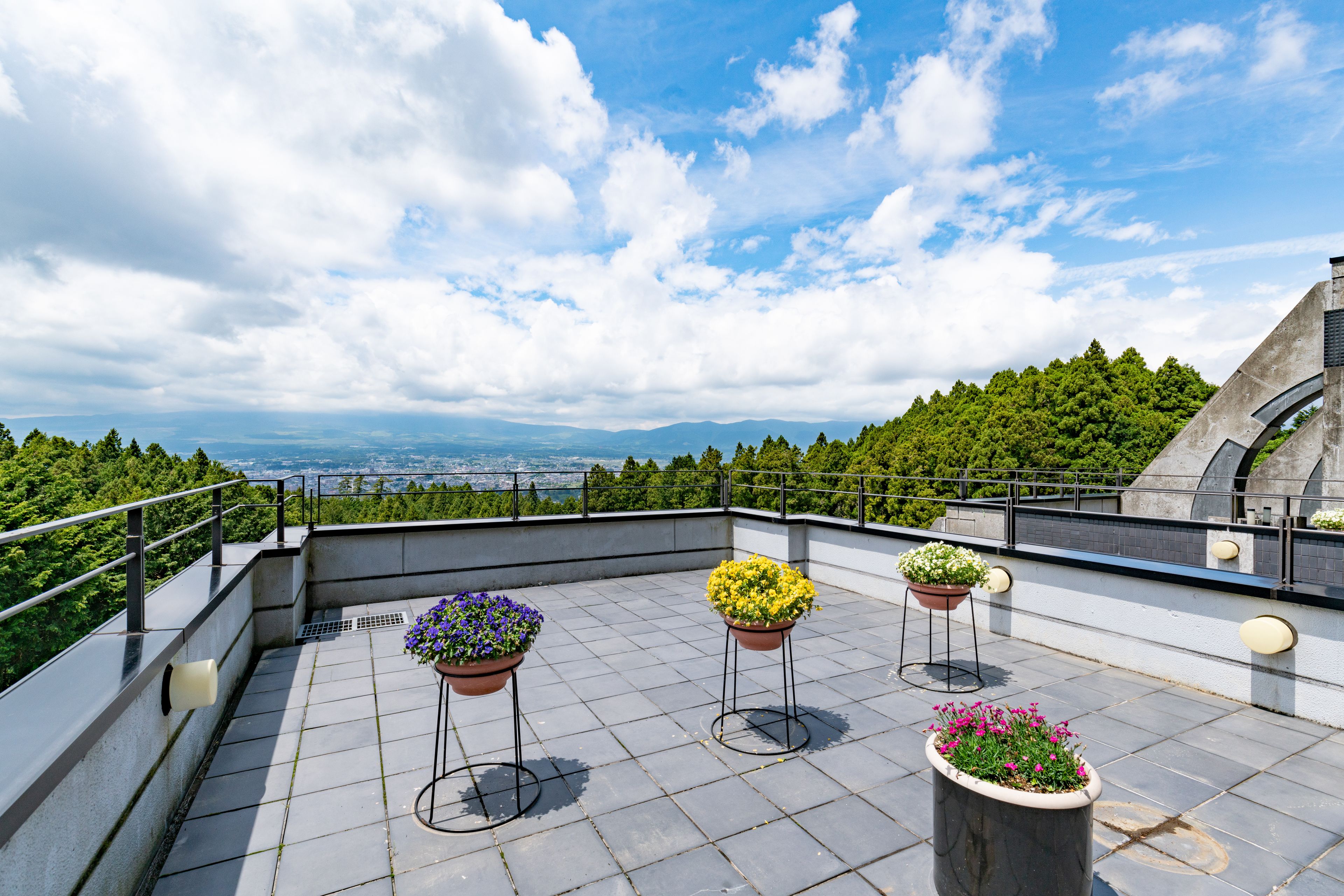 A rooftop terrace with three flower planters, a railing, and a view of a city and mountains.