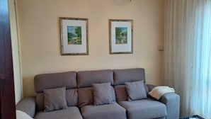 Una sala de estar con un sofá gris, dos cuadros en la pared, una mesa de centro de vidrio y una ventana con cortinas.