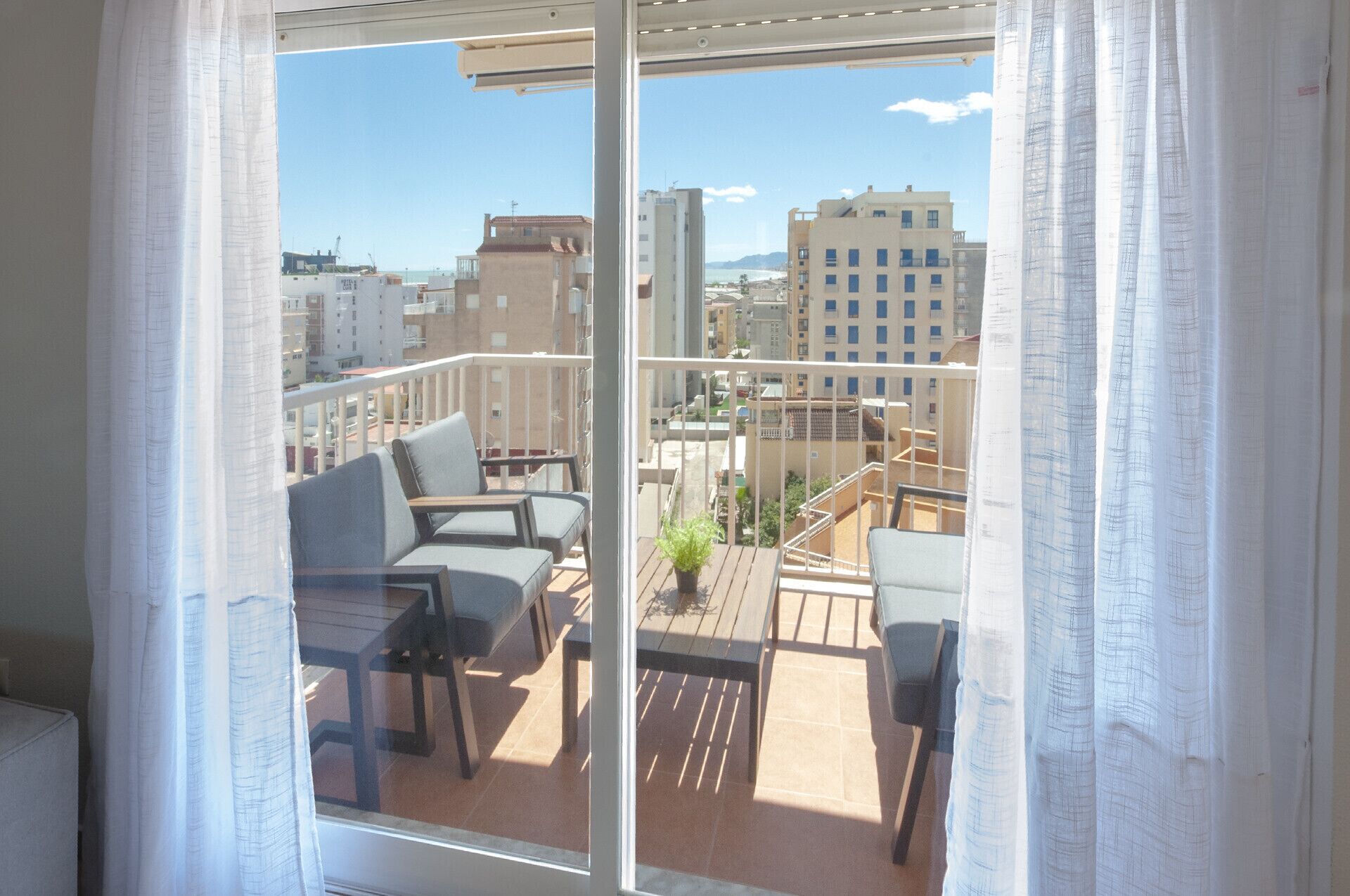 Un balcón con dos sillas y una mesita, con vistas a un paisaje urbano con edificios y cielo despejado.