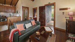 A cozy living room with a dark leather sofa, a wooden coffee table, and a kitchen area with wooden cabinets and a microwave. - Quinta Devónia - Cedro,country house with sea view (Funchal)