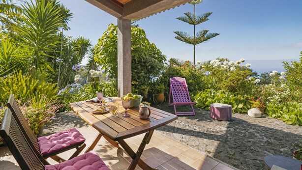 An outdoor patio with a wooden table and chairs, surrounded by lush greenery and a view of the ocean. - Quinta Devónia - Cedro,country house with sea view (Funchal)