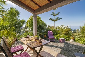 Um pátio externo com mesa e cadeiras de madeira, cercado por vegetação exuberante e vista para o oceano.