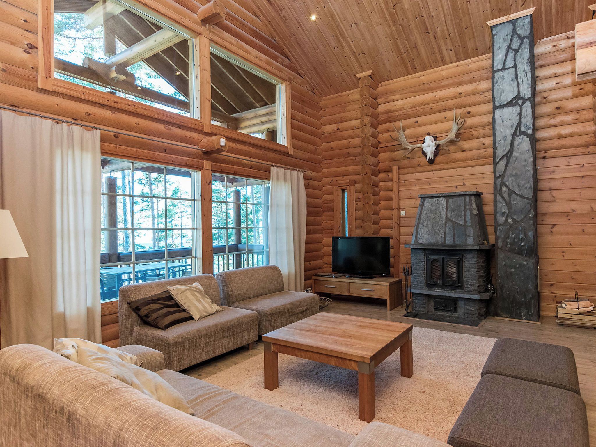 A log cabin interior with a sofa, a television, a fireplace, and large windows.