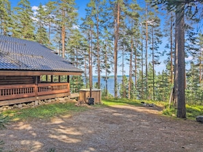 Eine Holzhütte mit einem Metalldach, umgeben von hohen Kiefern und einem Gewässer im Hintergrund.