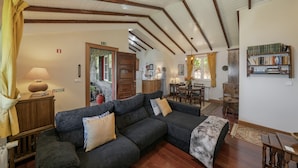 A cozy living room with a dark grey sofa, wooden beams on the ceiling, a bookshelf, and a dining area in the background. - Quinta Devónia - Tília,country house with sea view (Funchal)