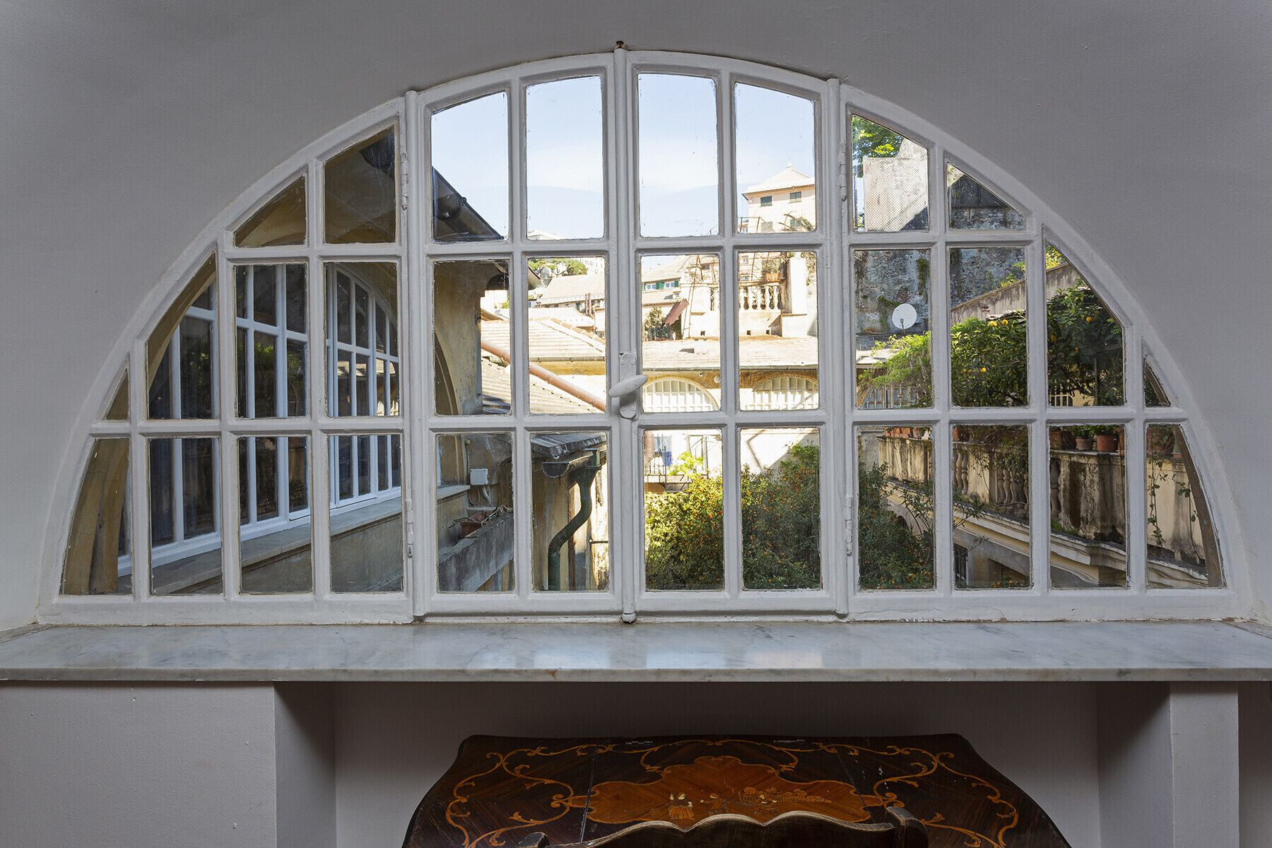 Une fenêtre à cadre blanc, composée de plusieurs vitres, donnant sur une cour intérieure avec des bâtiments et de la verdure.