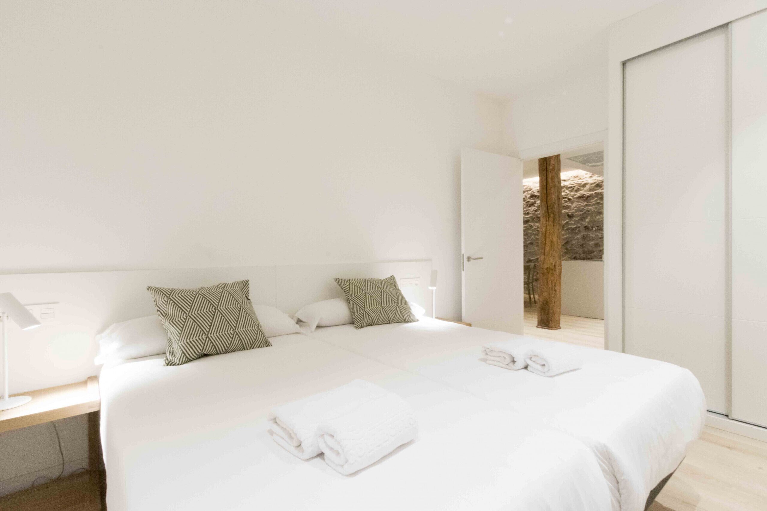 Quarto de hotel moderno com uma cama grande, roupa de cama branca e vista para uma parede de pedra por uma porta aberta.