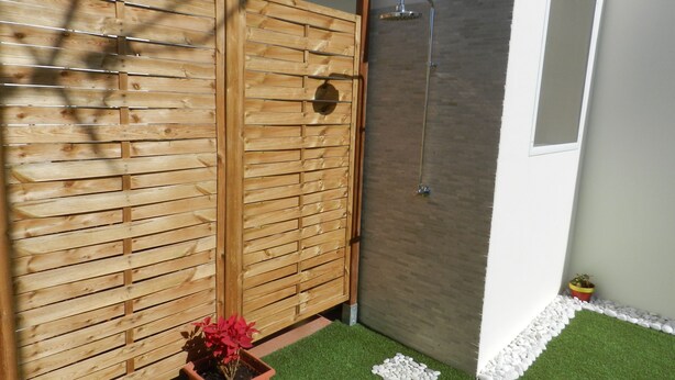 Une cloison en bois agrémentée d’un pot de plante, un espace douche avec un mur carrelé et une petite bordure de galets blancs.
