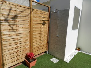 A wooden partition with a potted plant, a shower area with a tiled wall, and a small white pebbles border. - TROTAMUNDOS GRAN CANARIA 2 (El Tablero)