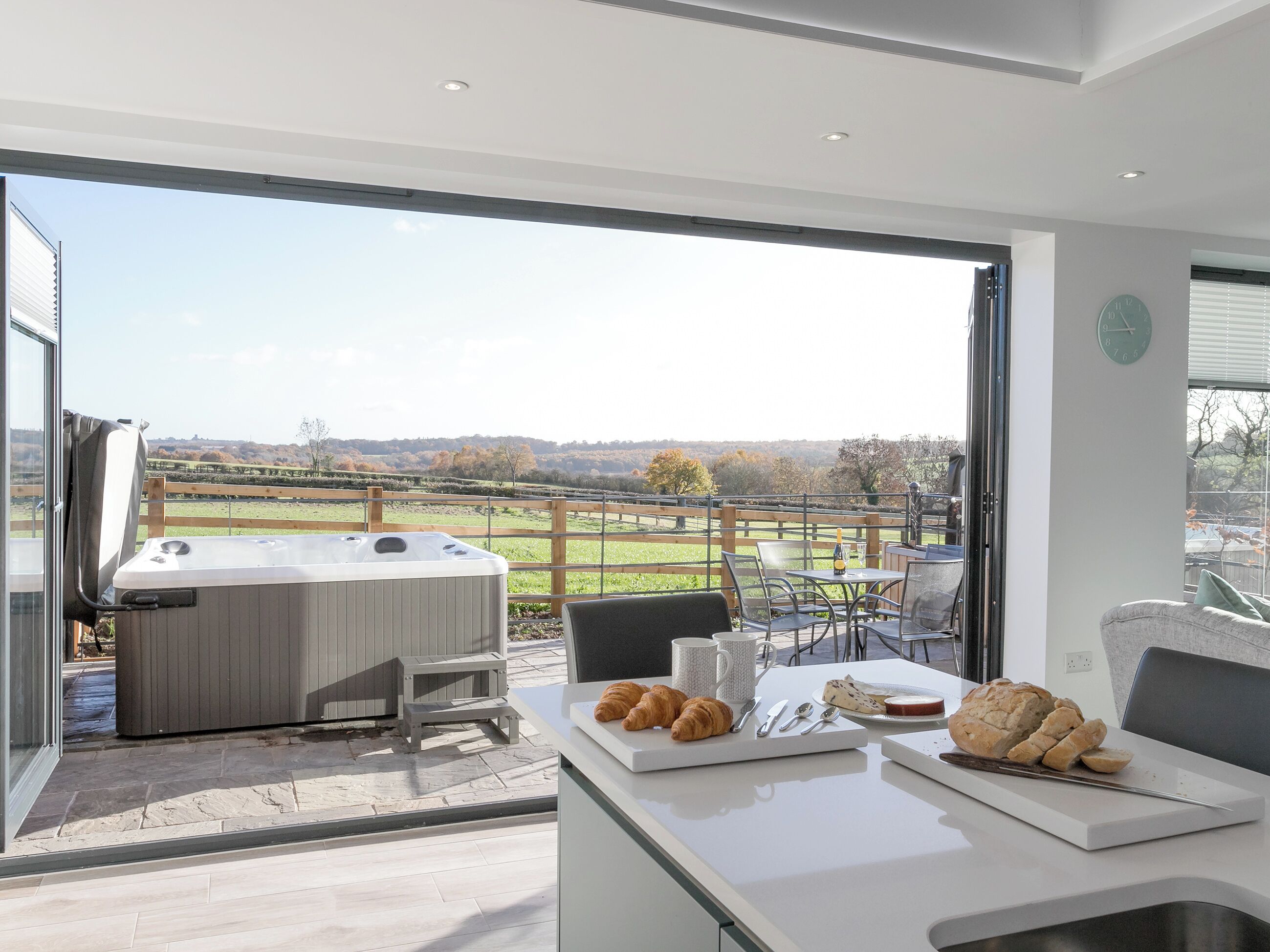 A modern kitchen with a large window overlooking a scenic countryside view, a hot tub, and a breakfast setup with croissants and cheese.
