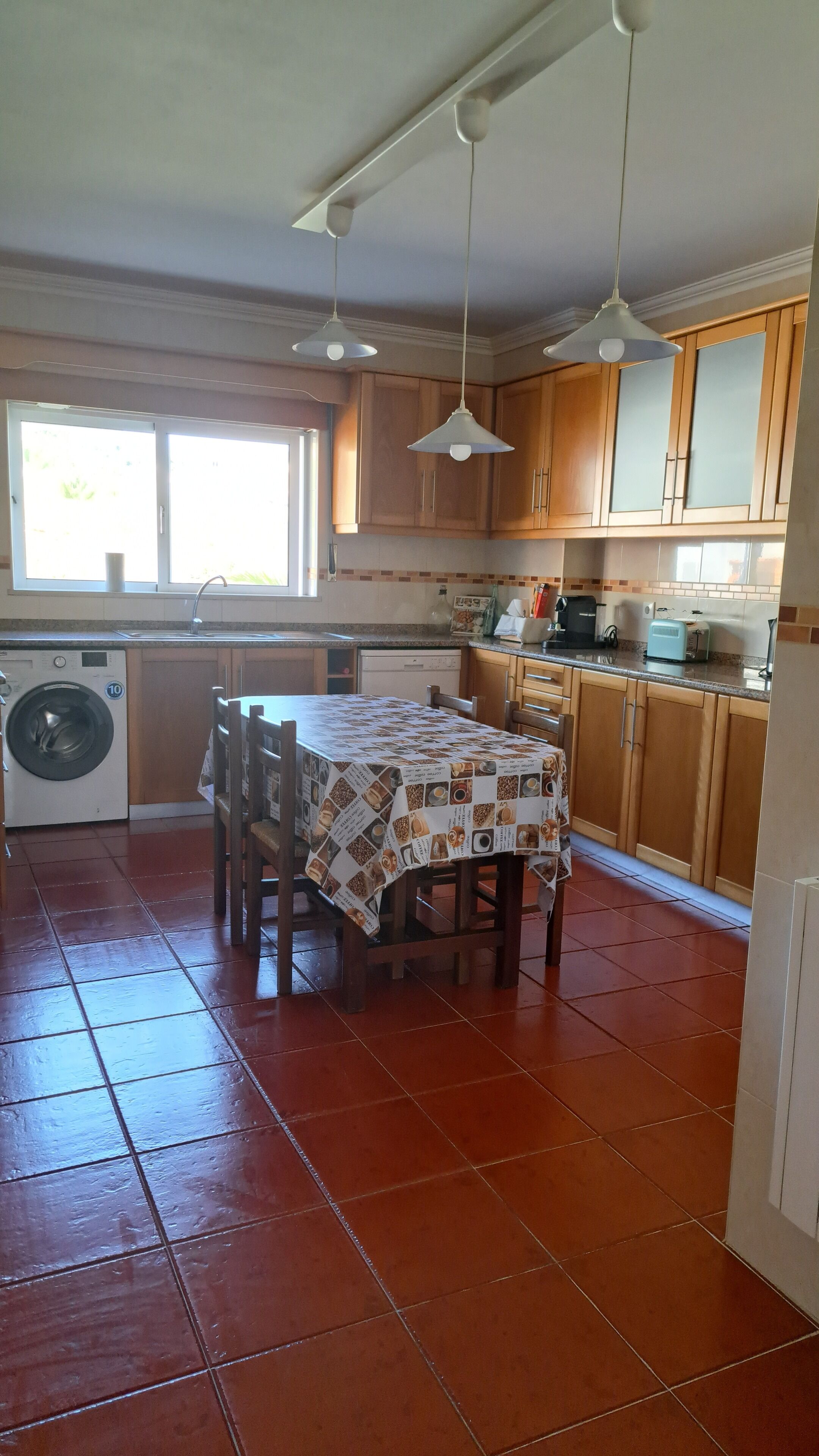 A kitchen with wooden cabinets, a washing machine, a table with a patterned tablecloth, and hanging pendant lights.