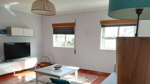 Un salon avec un téléviseur, des meubles blancs, une table à manger avec un saladier, un canapé avec des coussins turquoise et une colonne en bois.