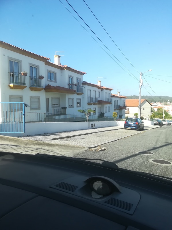 A row of white buildings with red-tiled roofs, balconies, and a paved street in the foreground.