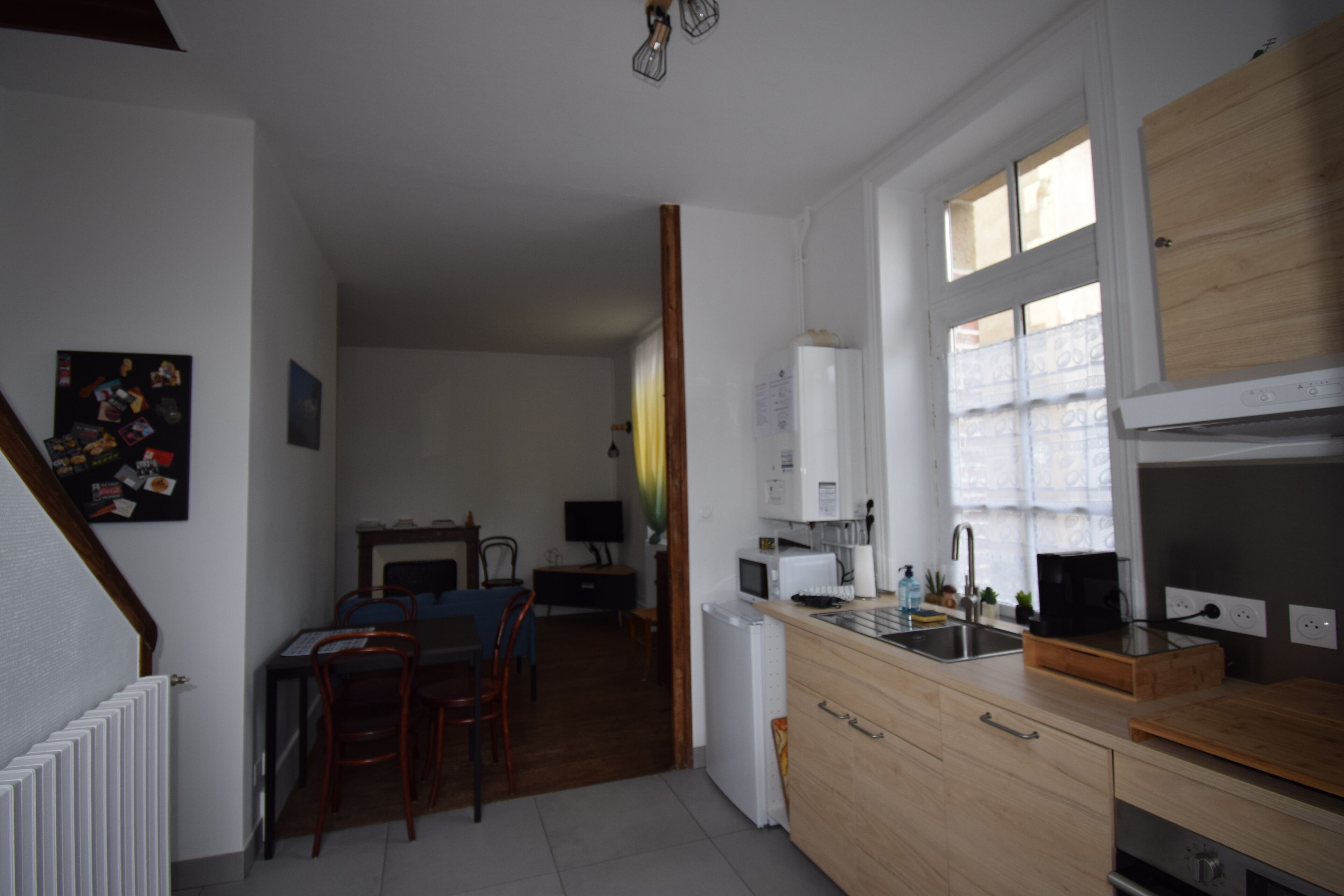 Une cuisine compacte avec des meubles en bois, un four à micro-ondes et un évier. On y trouve un coin repas avec des chaises et une table, et un réfrigérateur à l’arrière-plan.