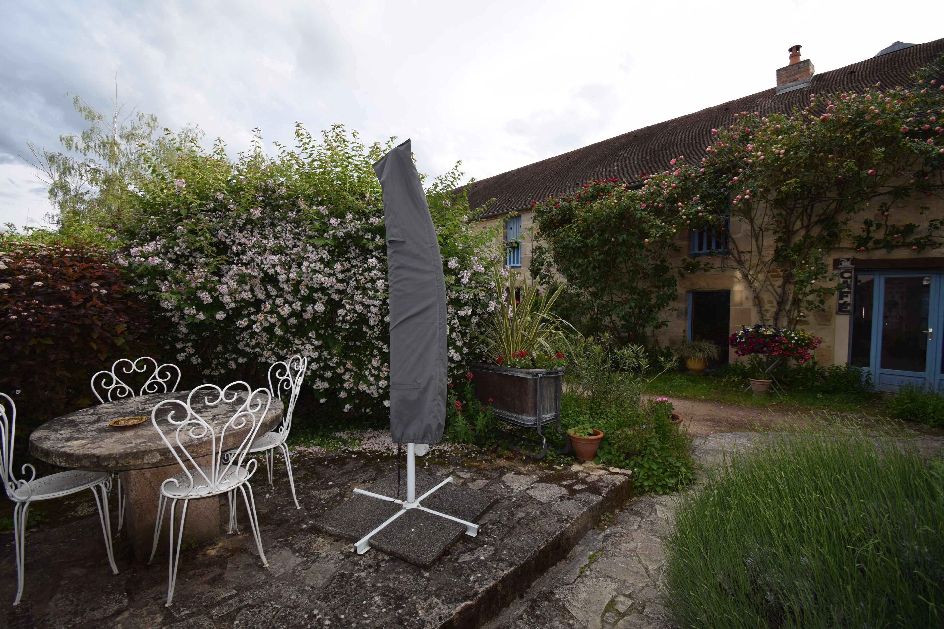 Un espace extérieur aménagé avec une table ronde et des chaises blanches, un parasol gris et une jardinière ornée de fleurs roses.