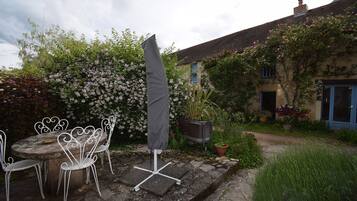 Un espace extérieur aménagé avec une table ronde et des chaises blanches, un parasol gris et une jardinière ornée de fleurs roses.