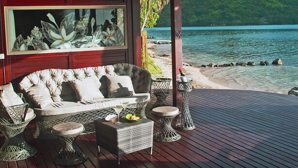 A covered outdoor seating area with a sofa, chairs, and tables, overlooking a beach and water.