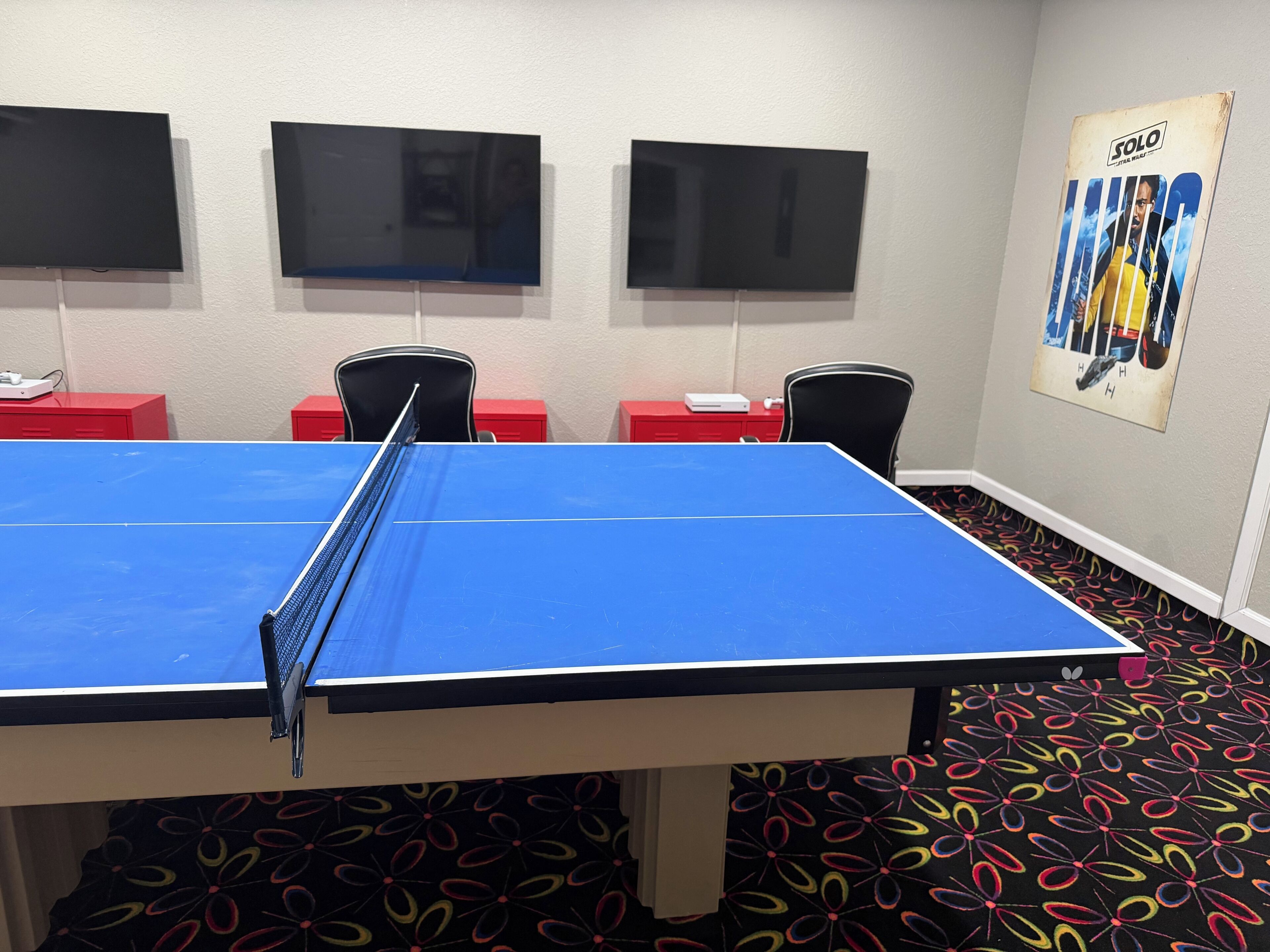 A ping pong table in a room with three flat-screen TVs mounted on the wall, red furniture, and a poster on the wall.