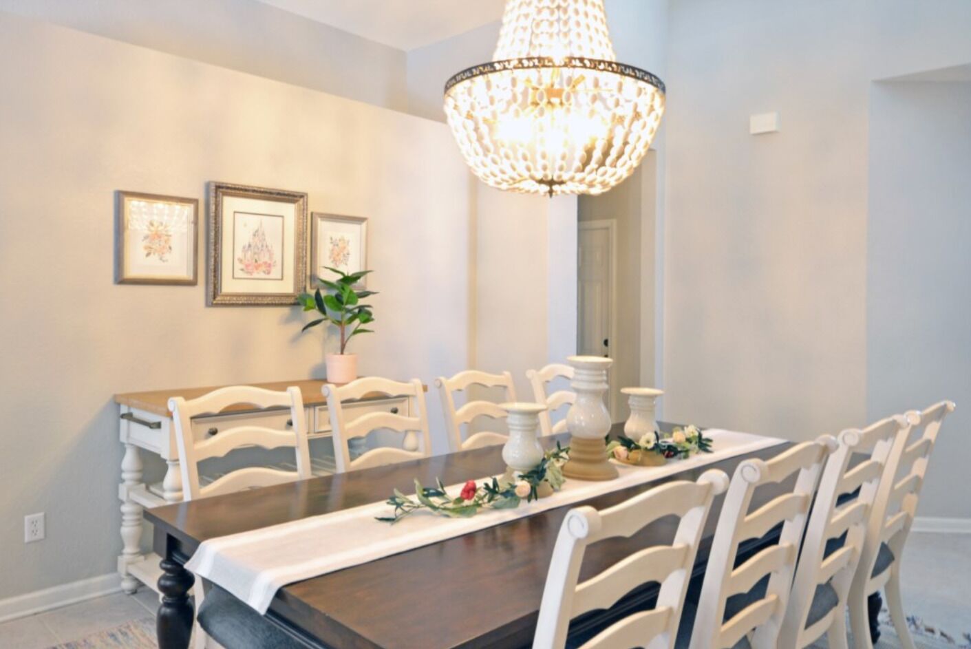Comedor con mesa de madera, sillas blancas, candelabro y cuadros en la pared.
