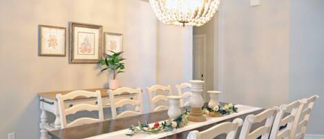 Comedor con mesa de madera, sillas blancas, candelabro y cuadros en la pared.