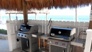 Two barbecue grills under a thatched roof, with a view of the ocean in the background.