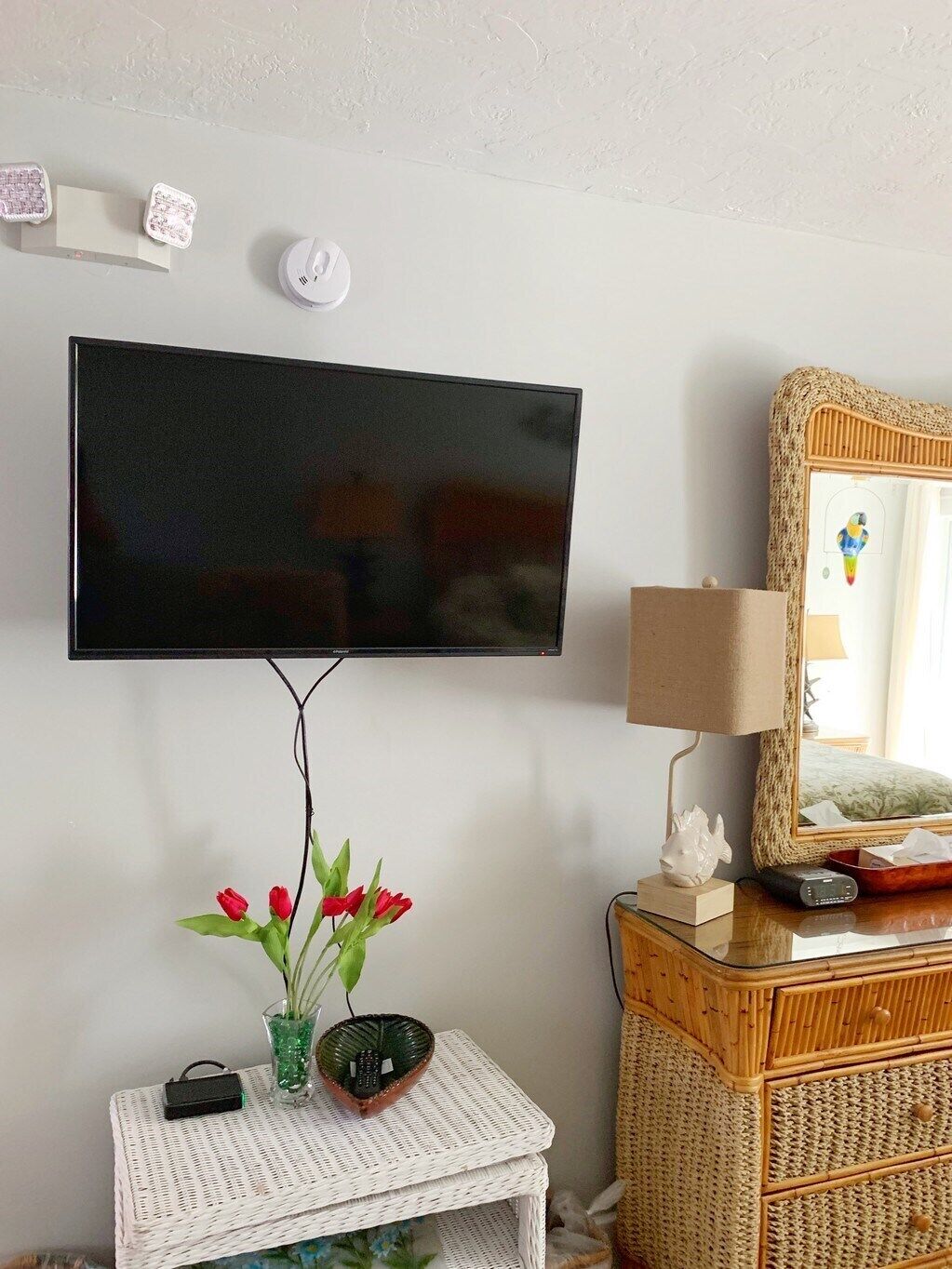 Un televisor de pantalla plana montado en la pared, una mesita de mimbre con un jarrón de tulipanes rojos, una lámpara y un espejo con marco de fibras.