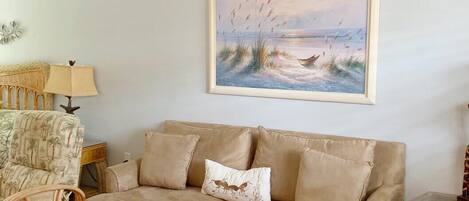 Un salon avec un canapé beige, une table basse en verre et un tableau représentant une scène de plage.