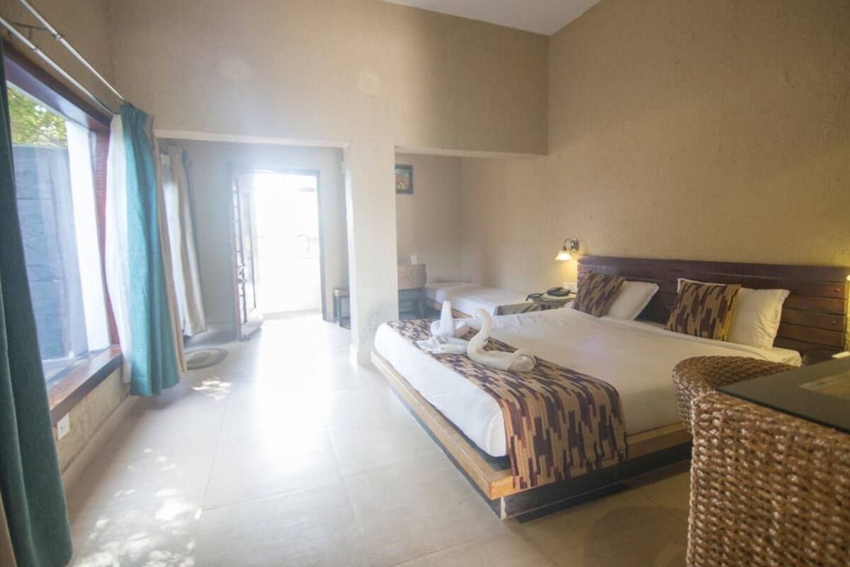 Habitación de hotel con una cama grande, una cama más pequeña, una silla de mimbre y un escritorio. Se ve una ventana con cortinas, una puerta que da a otra habitación y una lámpara fijada a la pared.