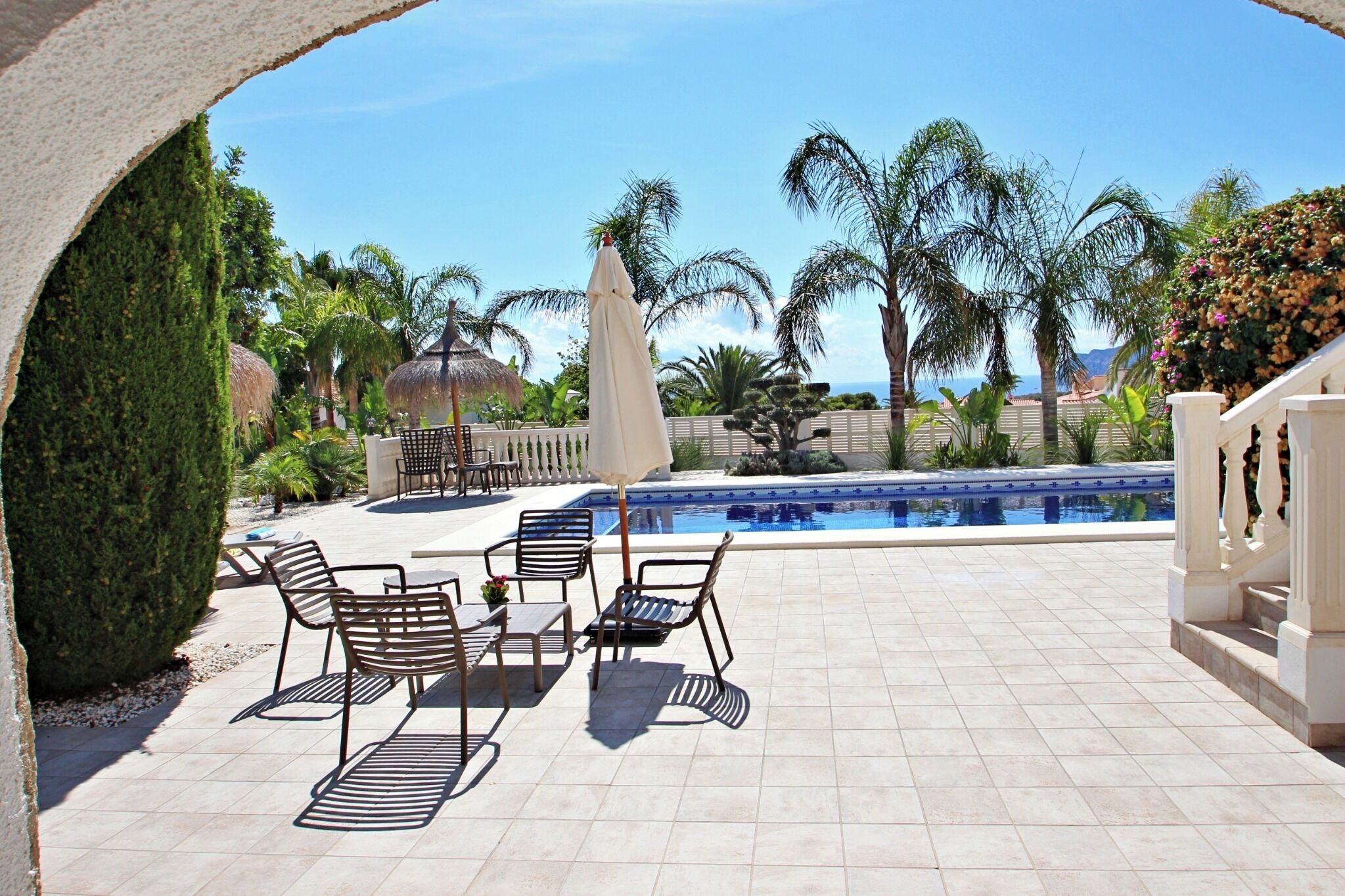 A poolside area with lounge chairs and a table, surrounded by palm trees and a view of the ocean.