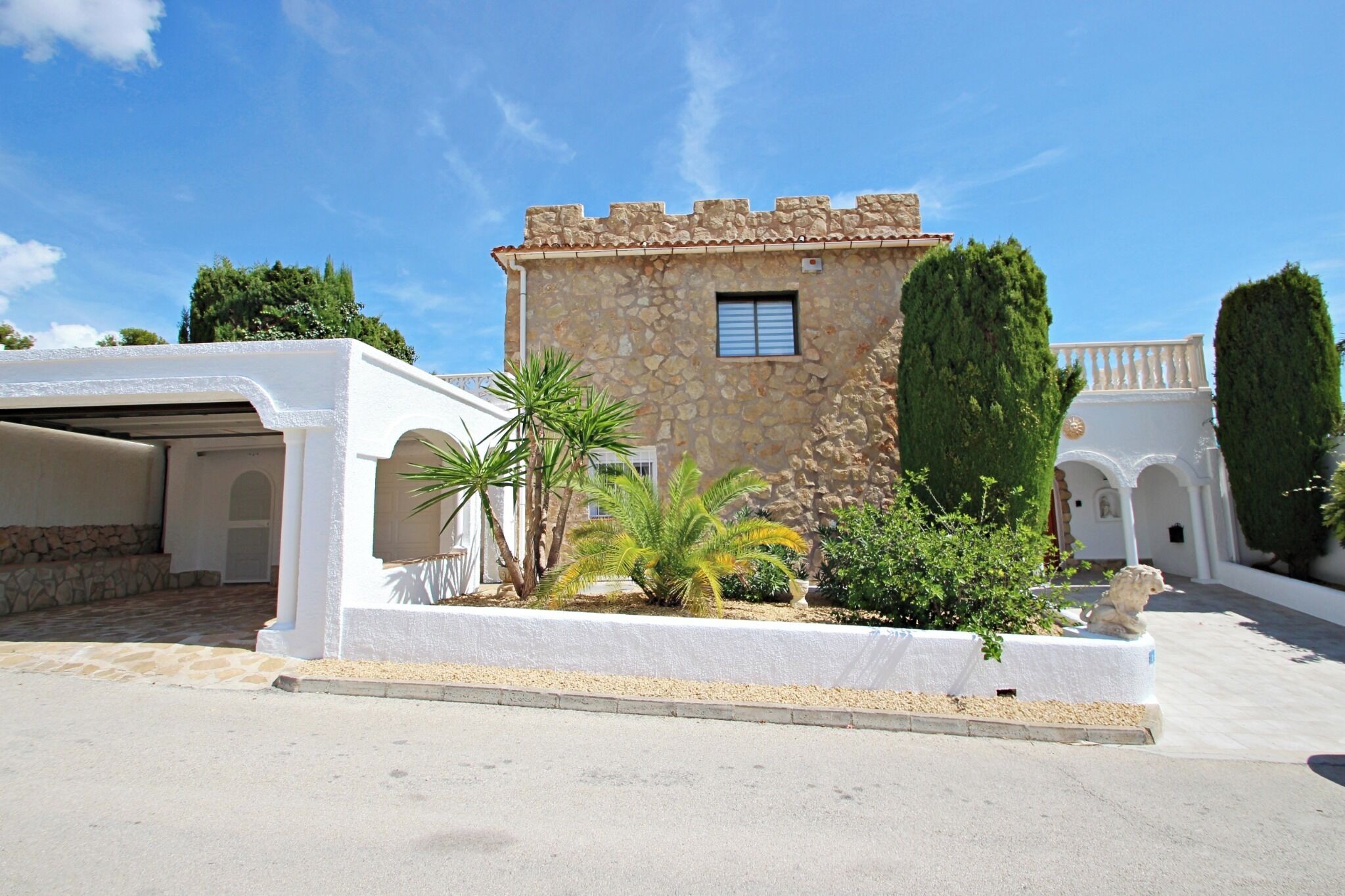 A stone building with a small window, surrounded by a white wall and a garden with palm trees and shrubs.